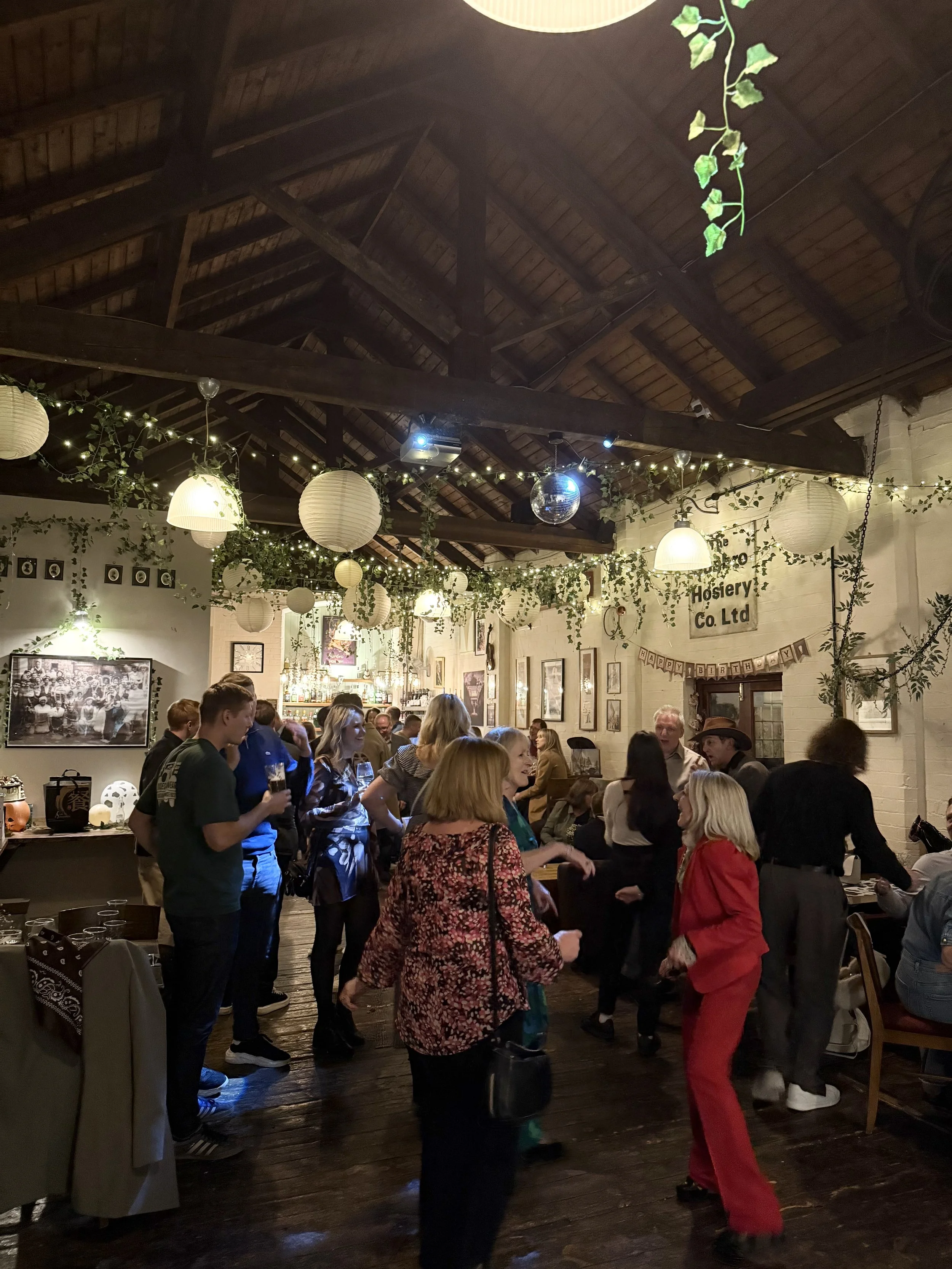 People socializing and dancing in a decorated indoor setting with hanging lanterns, fairy lights, and greenery.
