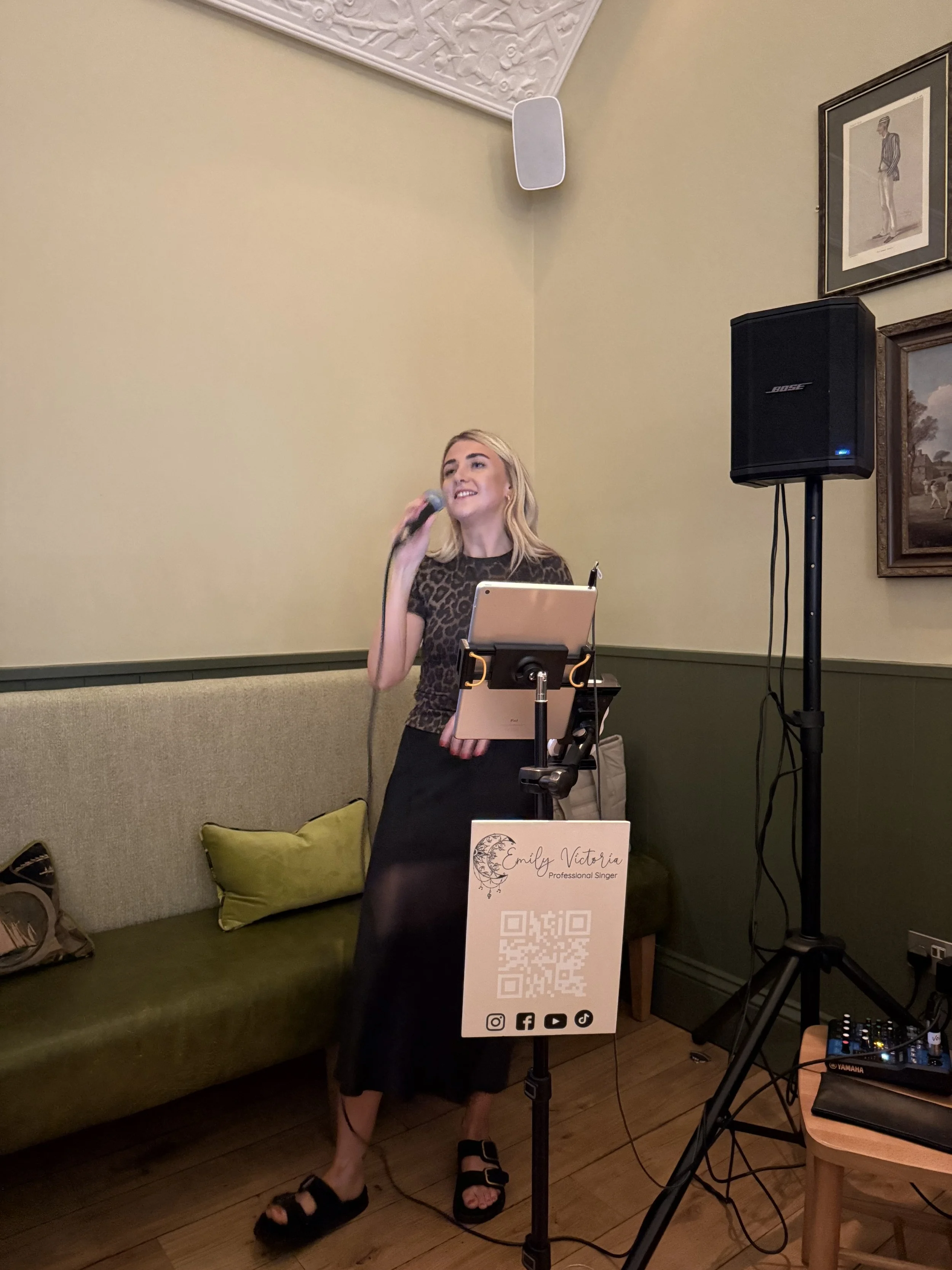 A woman singing into a microphone at an indoor event, standing in front of a green couch with two pillows, with a music stand displaying her name and social media icons, and a speaker and DJ equipment nearby.