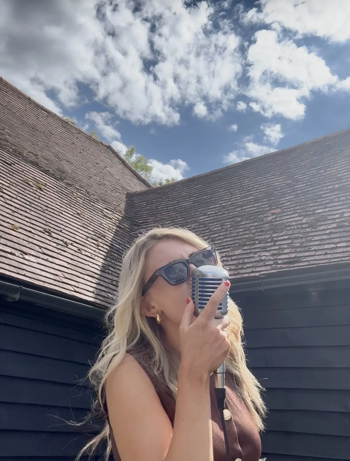 A woman with blonde hair, sunglasses, and gold earrings singing into a vintage microphone outdoors against a blue sky with clouds and rooftops.