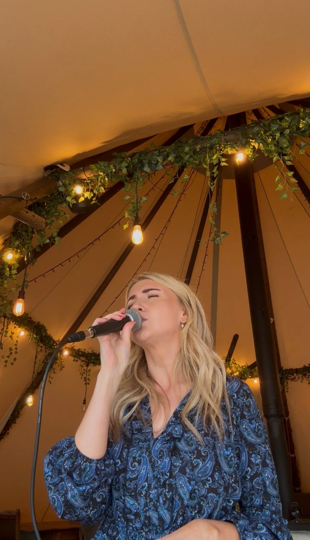 A blonde woman singing into a microphone inside a tent decorated with hanging string lights and greenery.
