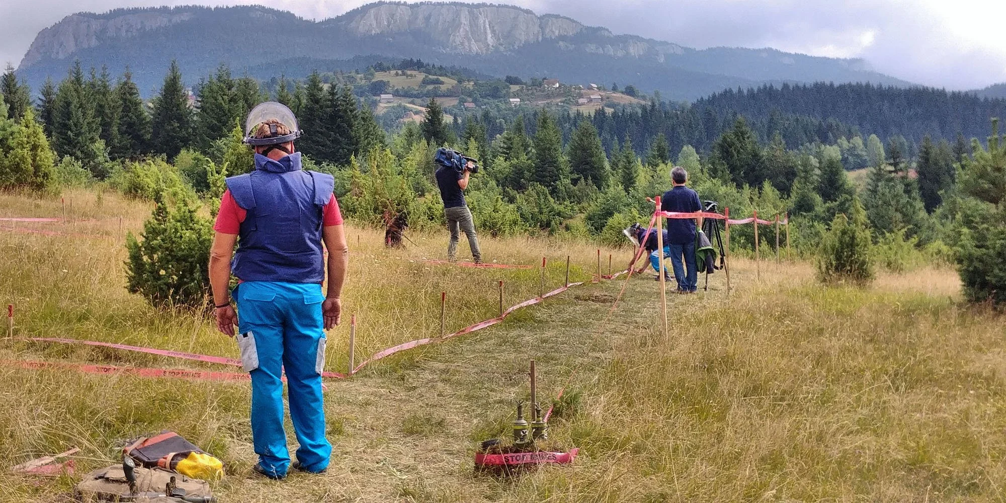 People working outdoors in a fenced grassy area with mountains and trees in the background. One person is filming with a camera, others are observing or working near equipment.