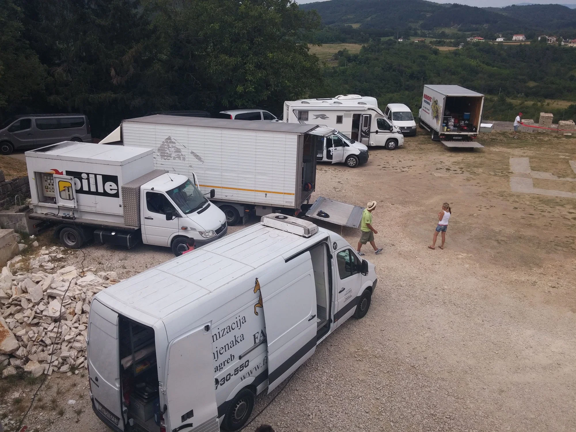 Several parked vehicles, including trucks, vans, and cars, on a gravel lot with some people walking around. There are trees and hills in the background.