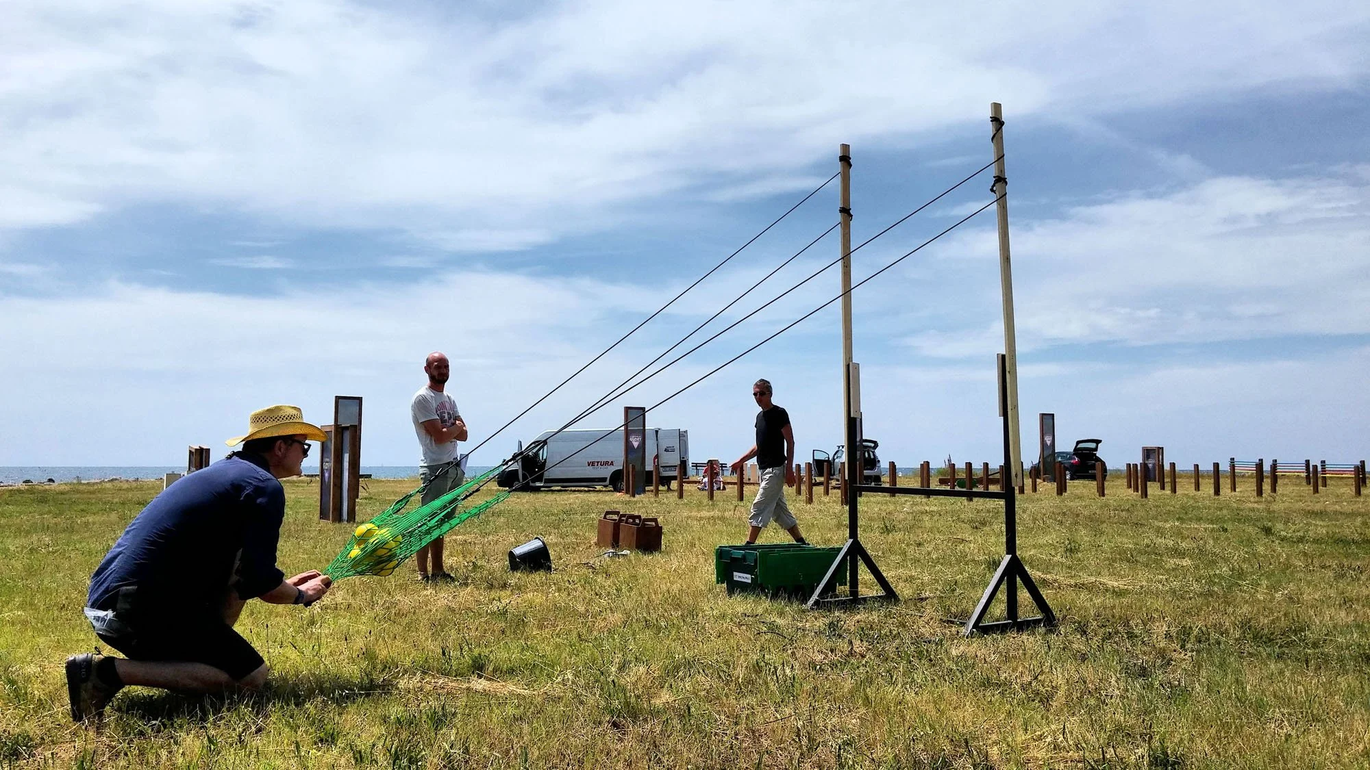 People setting up equipment outdoors on a grassy field near the ocean, with some standing and others kneeling, and a man wearing a straw hat.