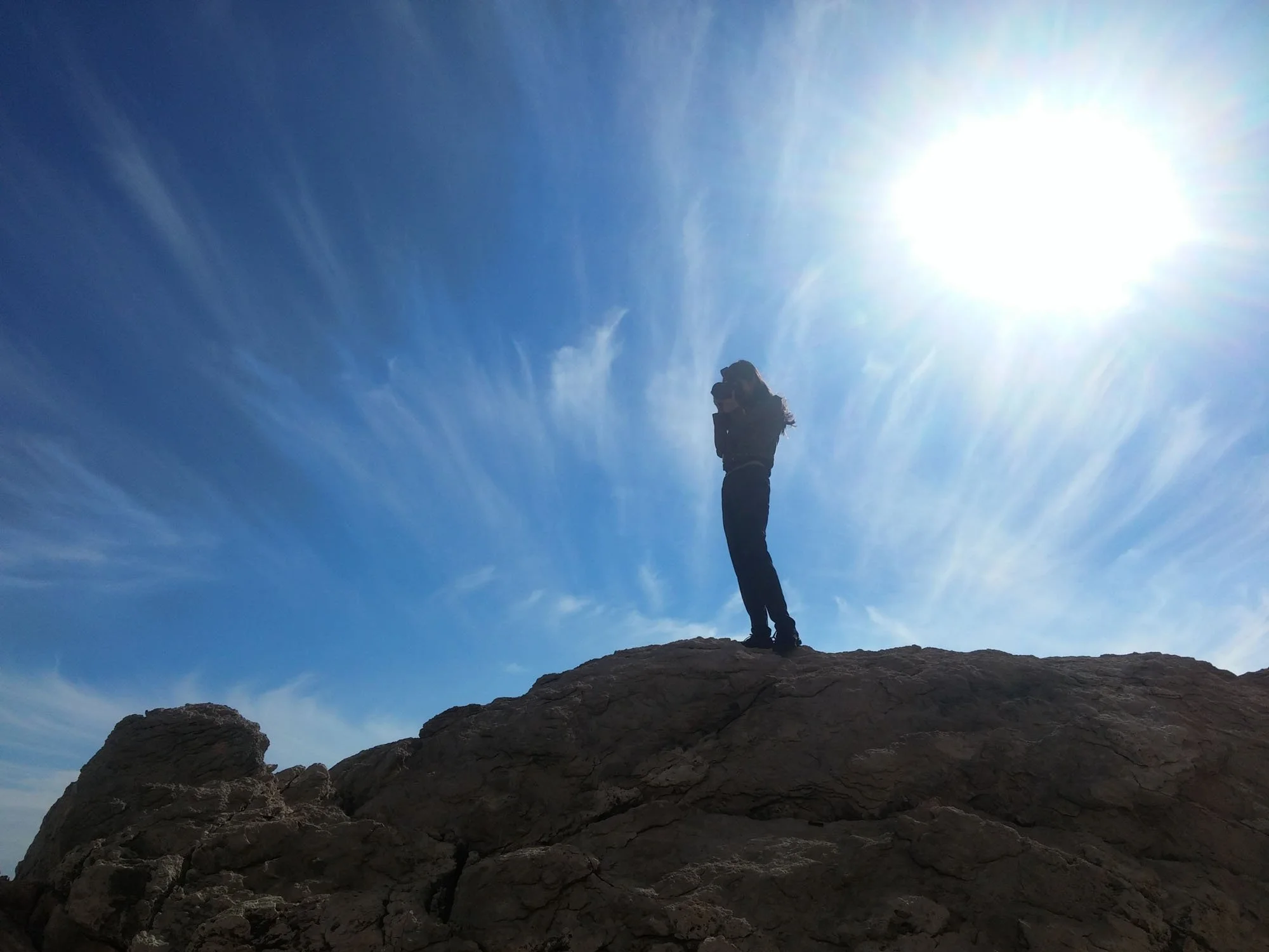 A person standing on a rocky mountaintop with arms crossed, silhouetted against a bright sun in a clear blue sky with wispy clouds.