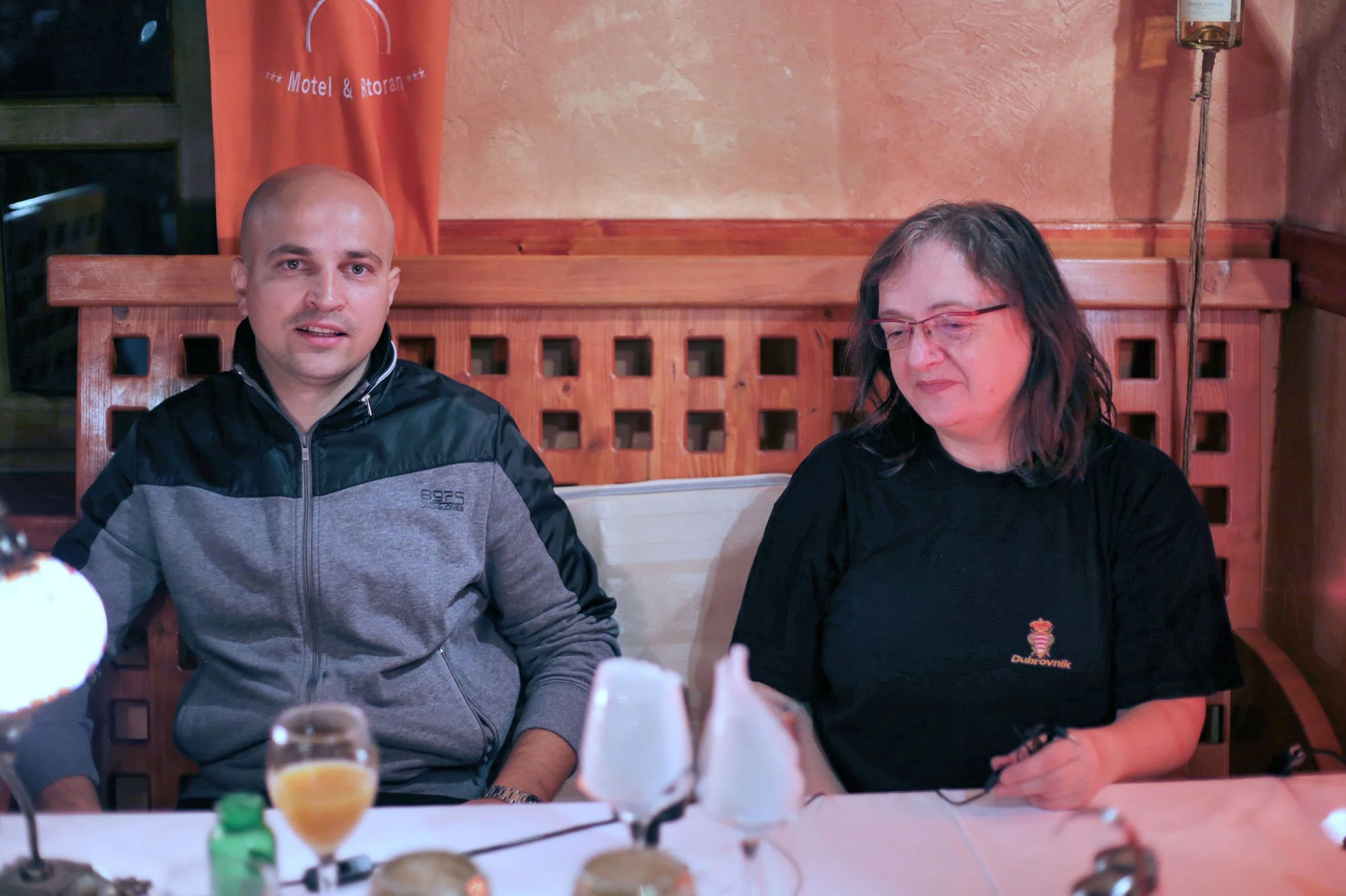 A man with a shaved head wearing a gray and black jacket sitting at a table with a woman with glasses and dark hair, wearing a black shirt. They are in a restaurant with warm-colored walls and wooden furniture.