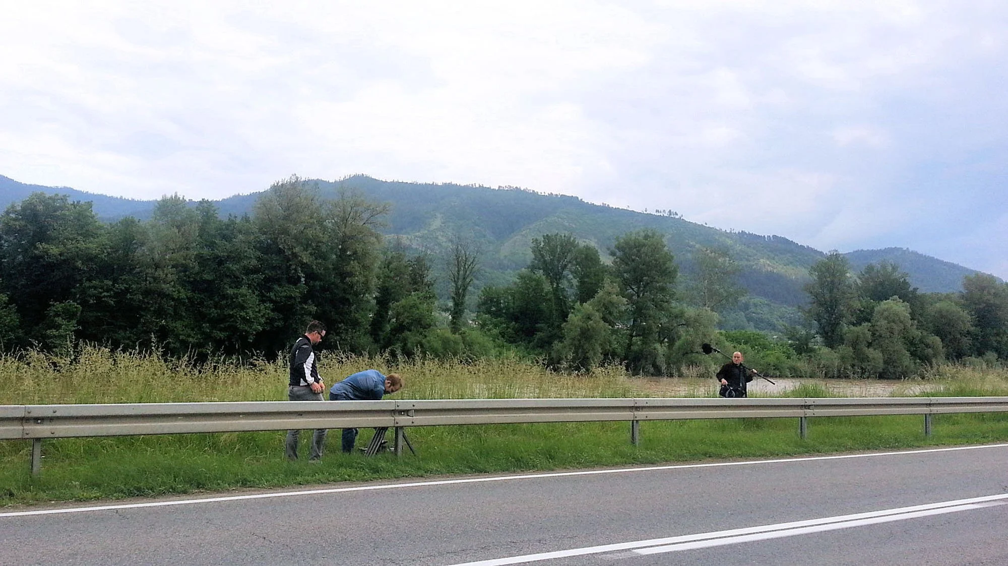 Four individuals near the side of a highway, with mountains and trees in the background. Three people are standing by a camera set up on a tripod, and one person is bending down, possibly adjusting equipment. The scene appears to involve filming or t