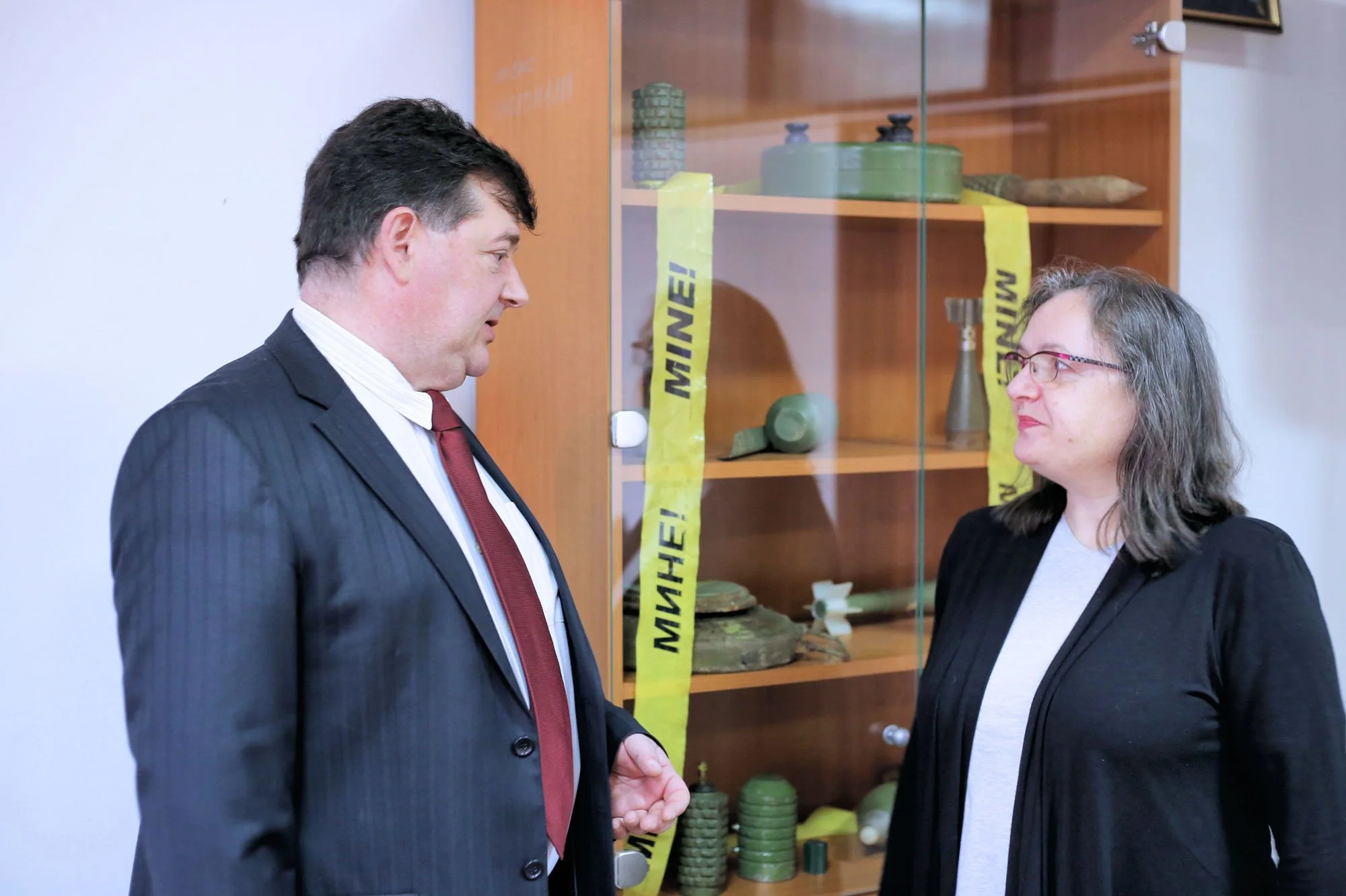 Two people standing and talking in front of a glass display case with military gear, with yellow caution tape on the case.