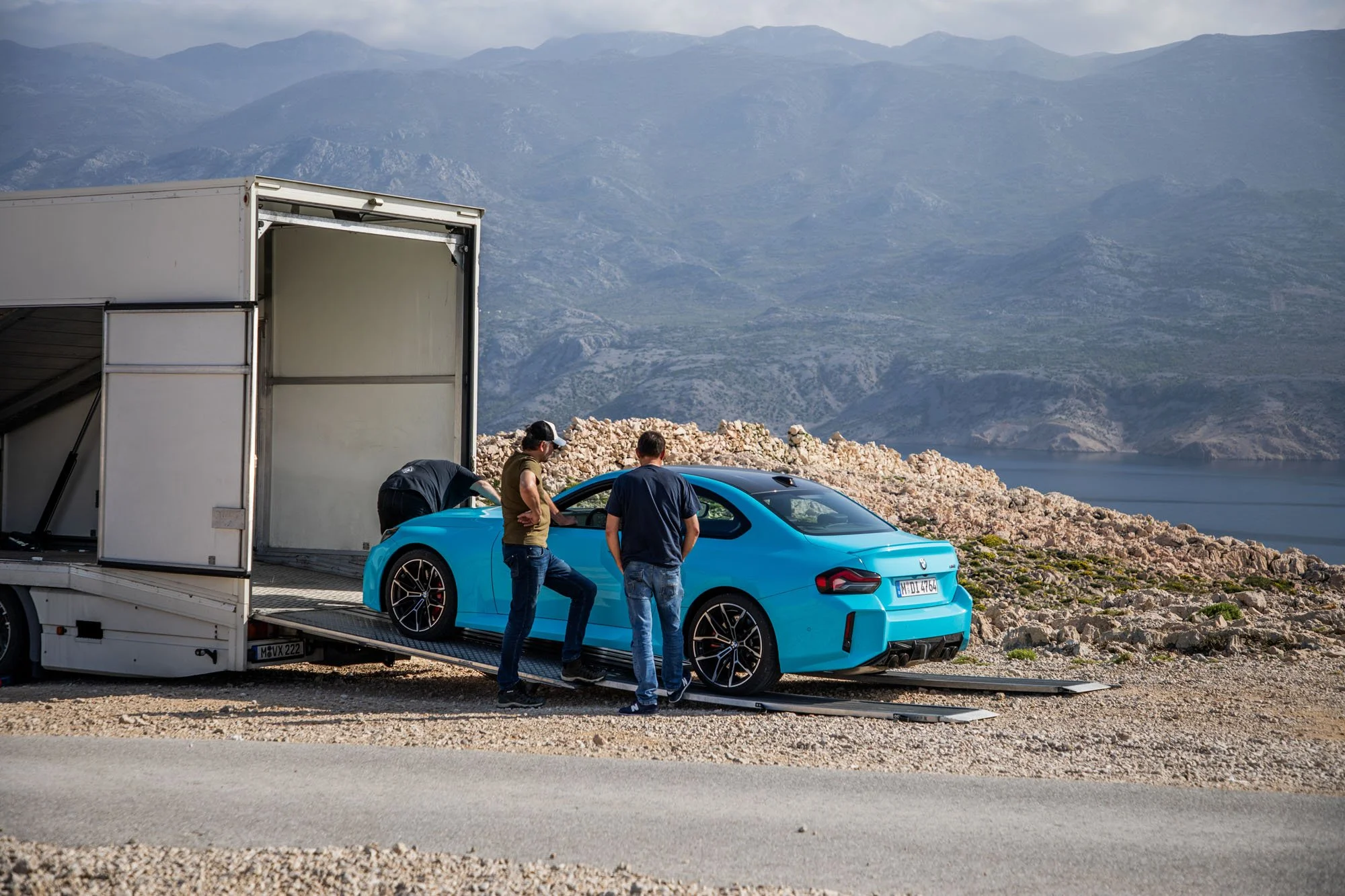 BMW M2 M xDrive production in Croatia as the car is unloaded from a transport truck, with the film crew, Pag Island, the Velebit mountains, and the Adriatic Sea in the background.