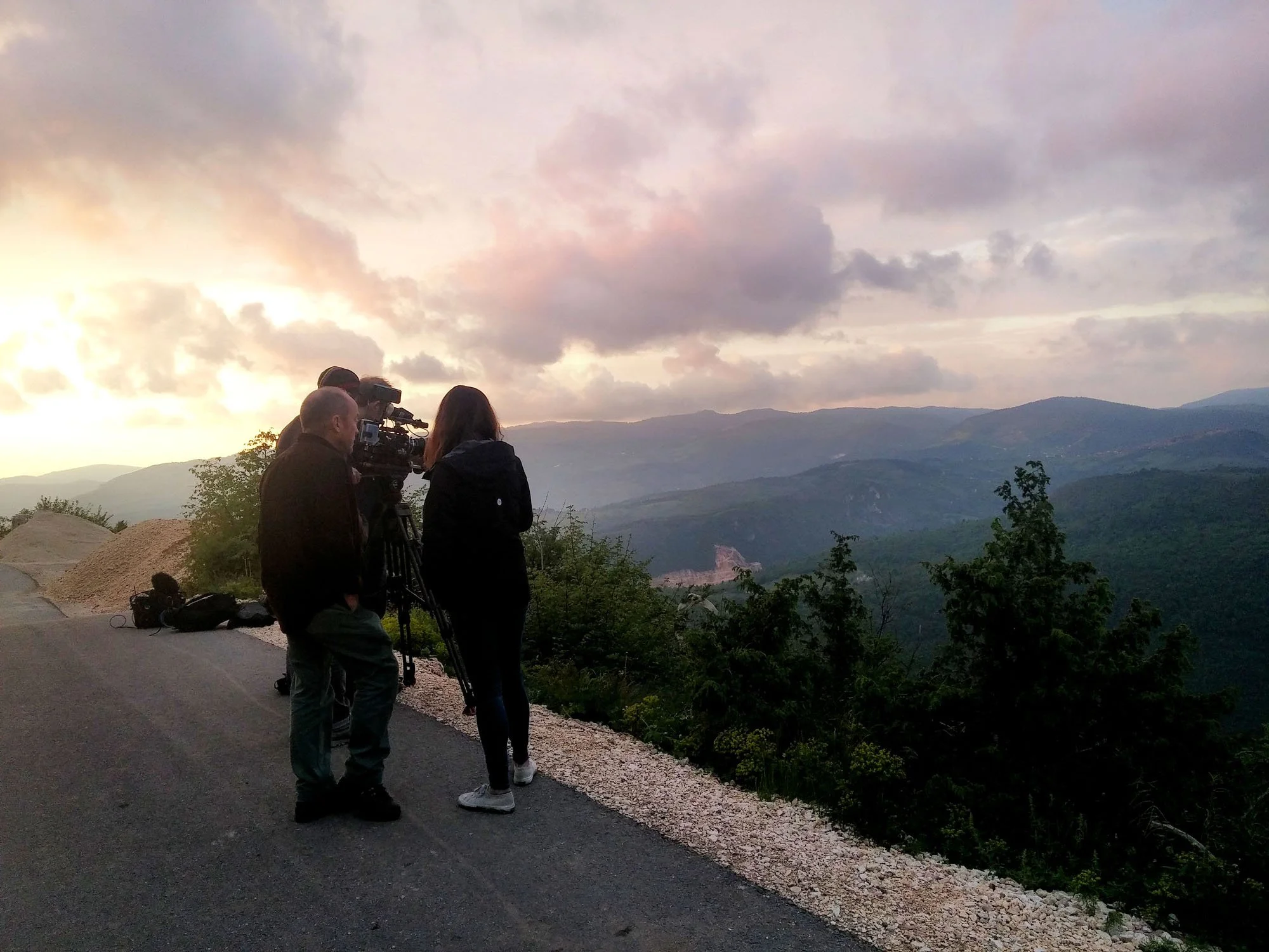 Two women and one man are standing by a camera on a tripod, capturing a sunset view of distant mountains and a cloudy sky at dusk.