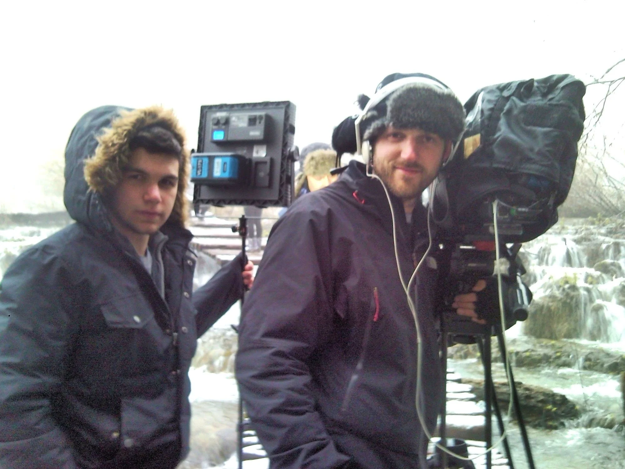 Two men are outdoors, dressed warmly with jackets, among trees and a waterfall. One is holding a camera on a tripod, and the other is wearing headphones and a large backpack.