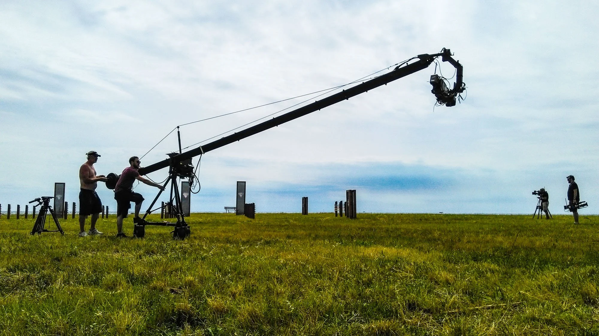 A film crew operating a camera crane outdoors in a grassy field on a cloudy day.