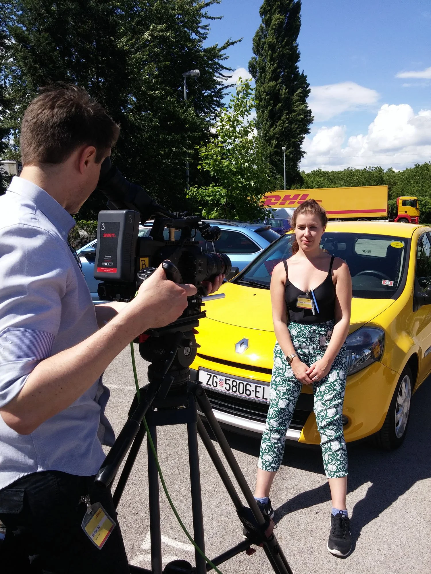 A woman standing in front of a yellow car being filmed by a man with a camera. The woman is wearing a black tank top and patterned pants, and is holding a clipboard. The setting appears to be a parking lot with trees and DHL trucks in the background.