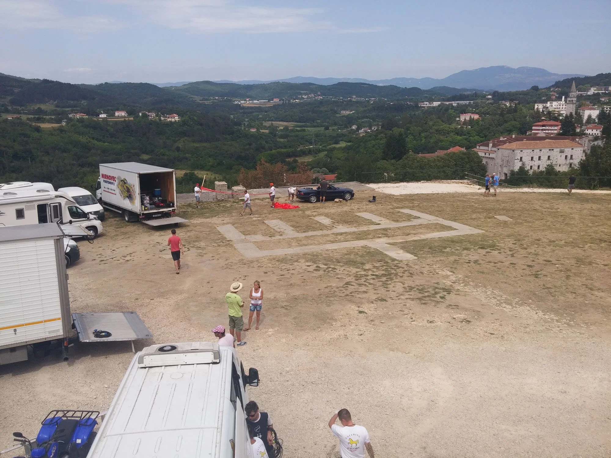 People setting up outdoor event equipment on a hilltop with a scenic view of distant mountains and a small town in the background.