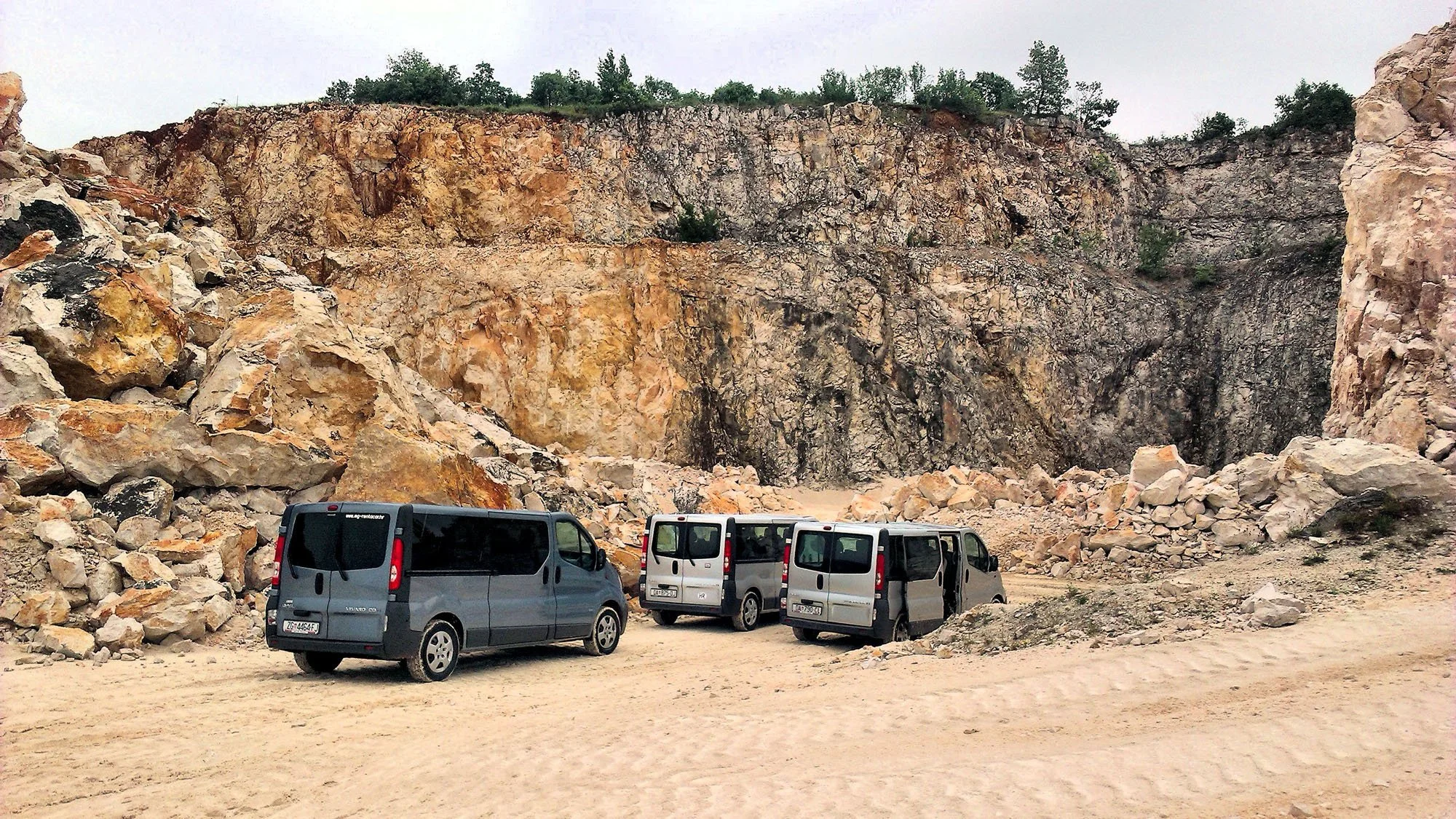 A rocky, mountainous area with a few small trees and a cluster of parked vans at the base of the cliffs.