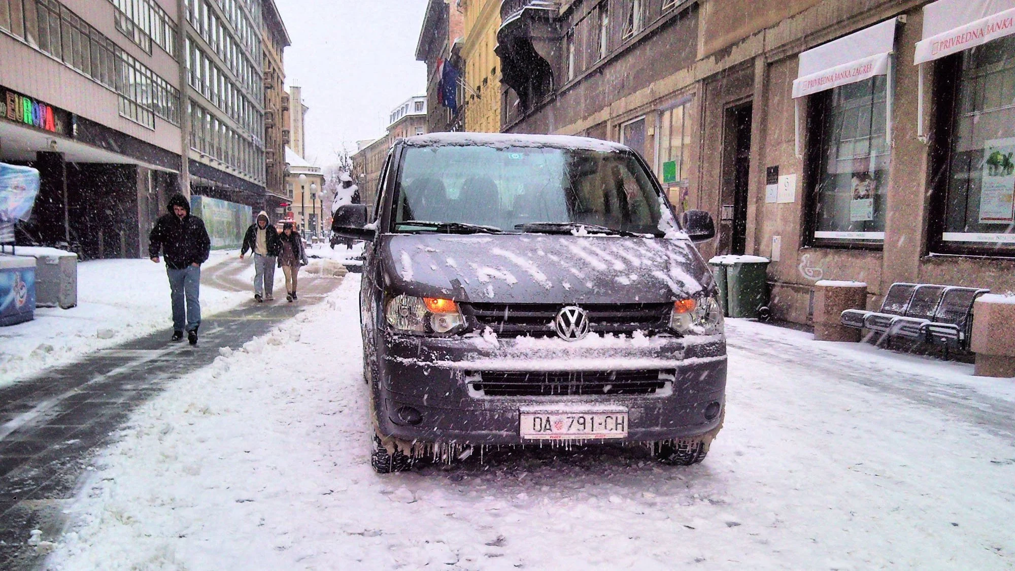 A black Volkswagen van covered in snow parked on a city street during a snowstorm, with pedestrians walking past and buildings on either side.