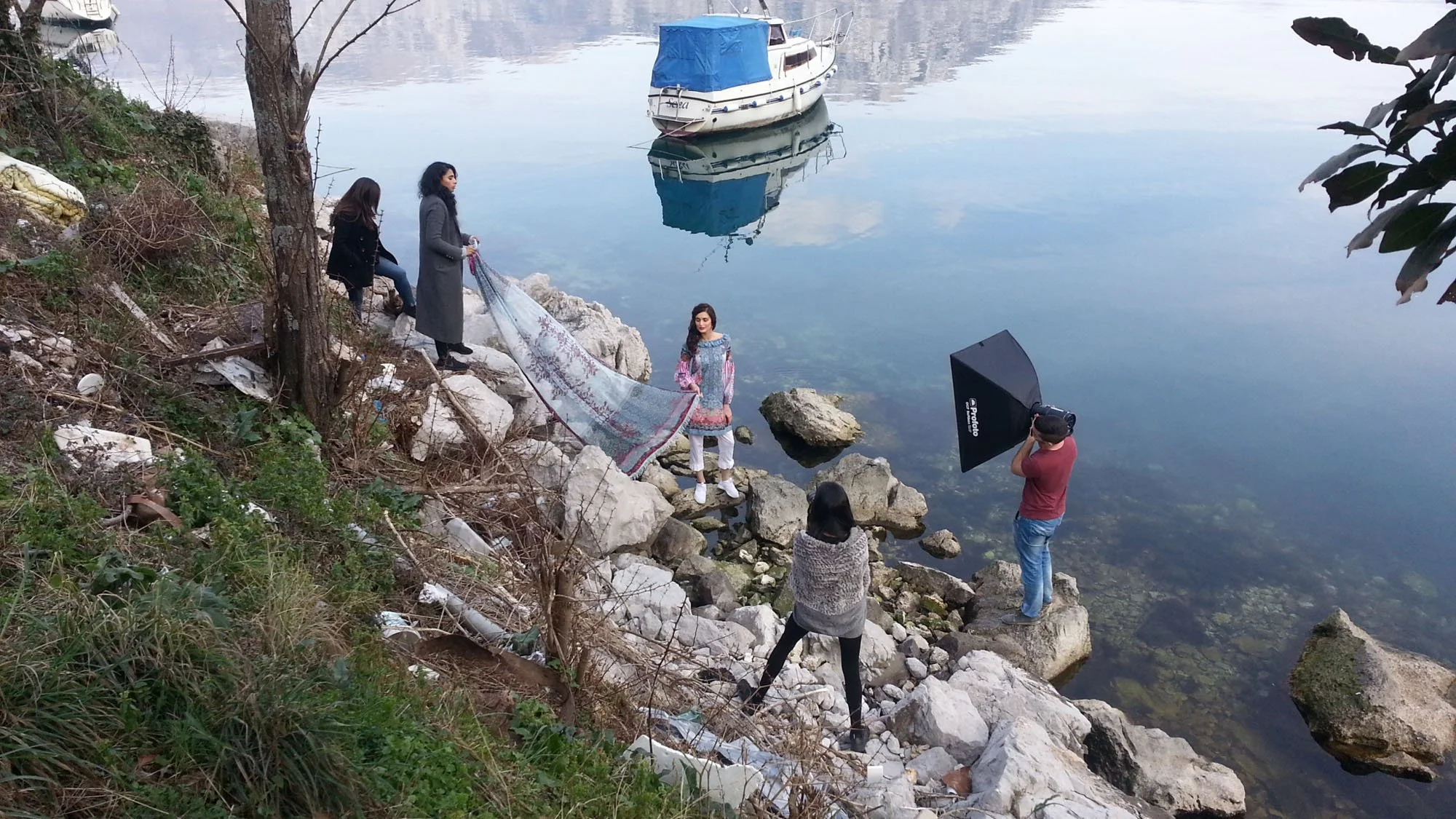 A photoshoot by the water showing a woman in a dress standing on rocks, two women with a rug on the rocks, and a photographer with a large umbrella, while two other women are sitting on rocks on the hillside.