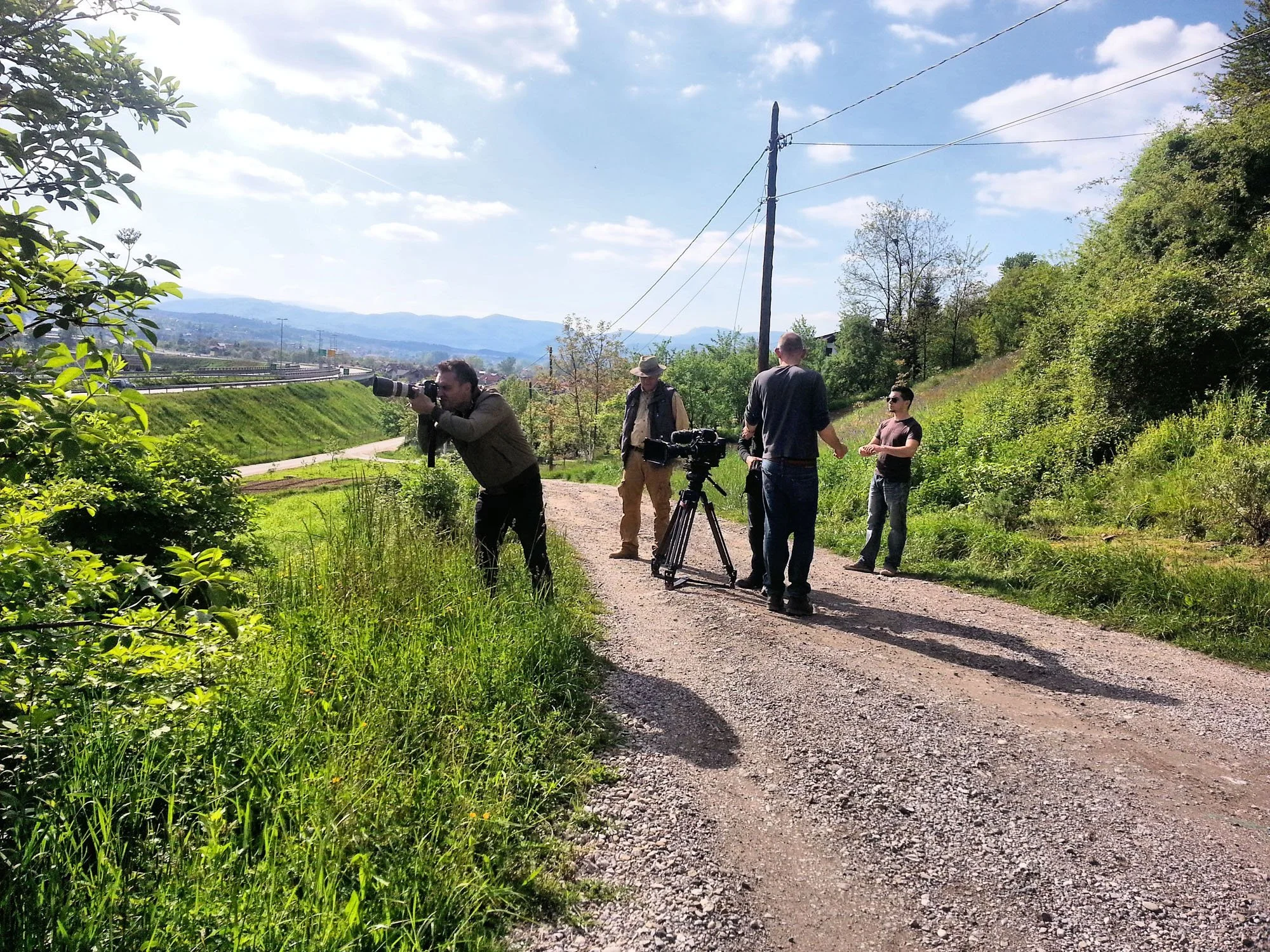 A group of five men with cameras and filming equipment gathered outdoors on a gravel path near greenery, with mountains and a partly cloudy sky in the background.