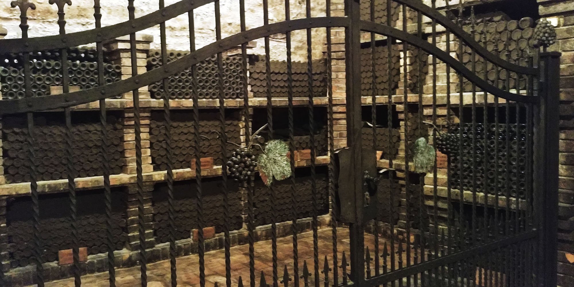 A cellar with stacks of wine bottles stored on shelves, visible through a black iron gate with decorative grapevine details and a locked gate handle.