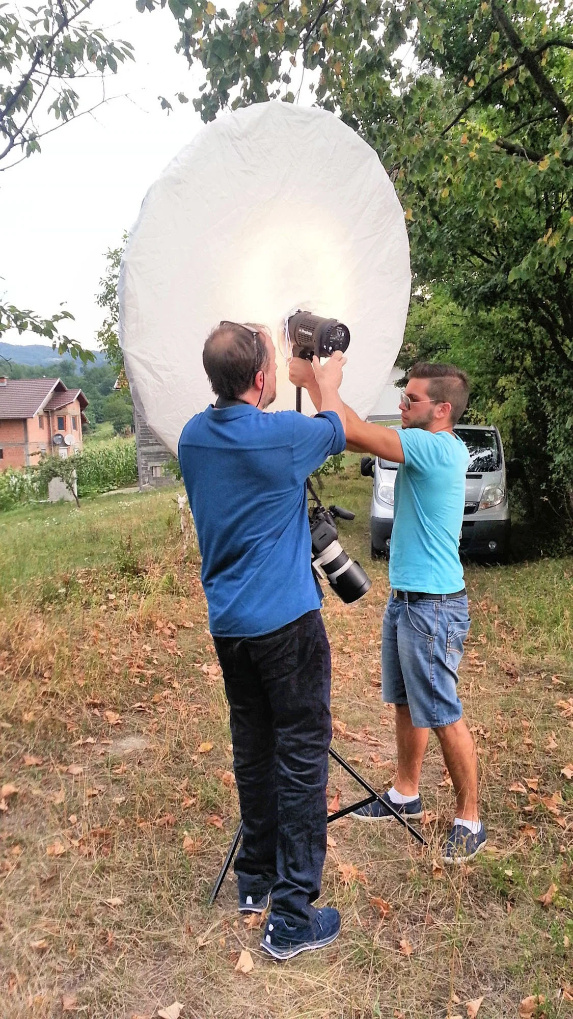 Two men are standing outdoors in a grassy area with trees and houses in the background. One man is holding a large white umbrella and adjusting a professional camera with a telephoto lens. The other man is assisting, wearing sunglasses and a light bl