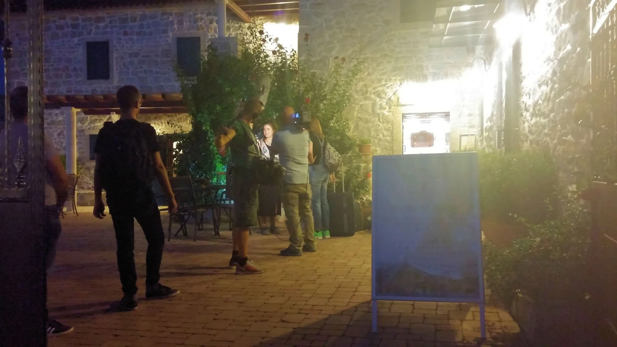 Group of people in an outdoor setting near a stone building, some with cameras, illuminated by bright lights, with a brick-paved floor and plants around.