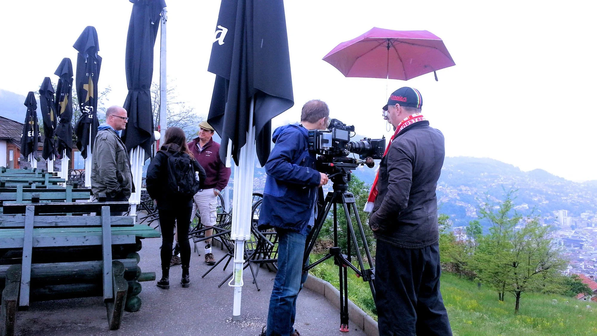 Group of people filming or taking photos outdoors on a rainy day, with some holding umbrellas, in a scenic location with trees and hills in the background.