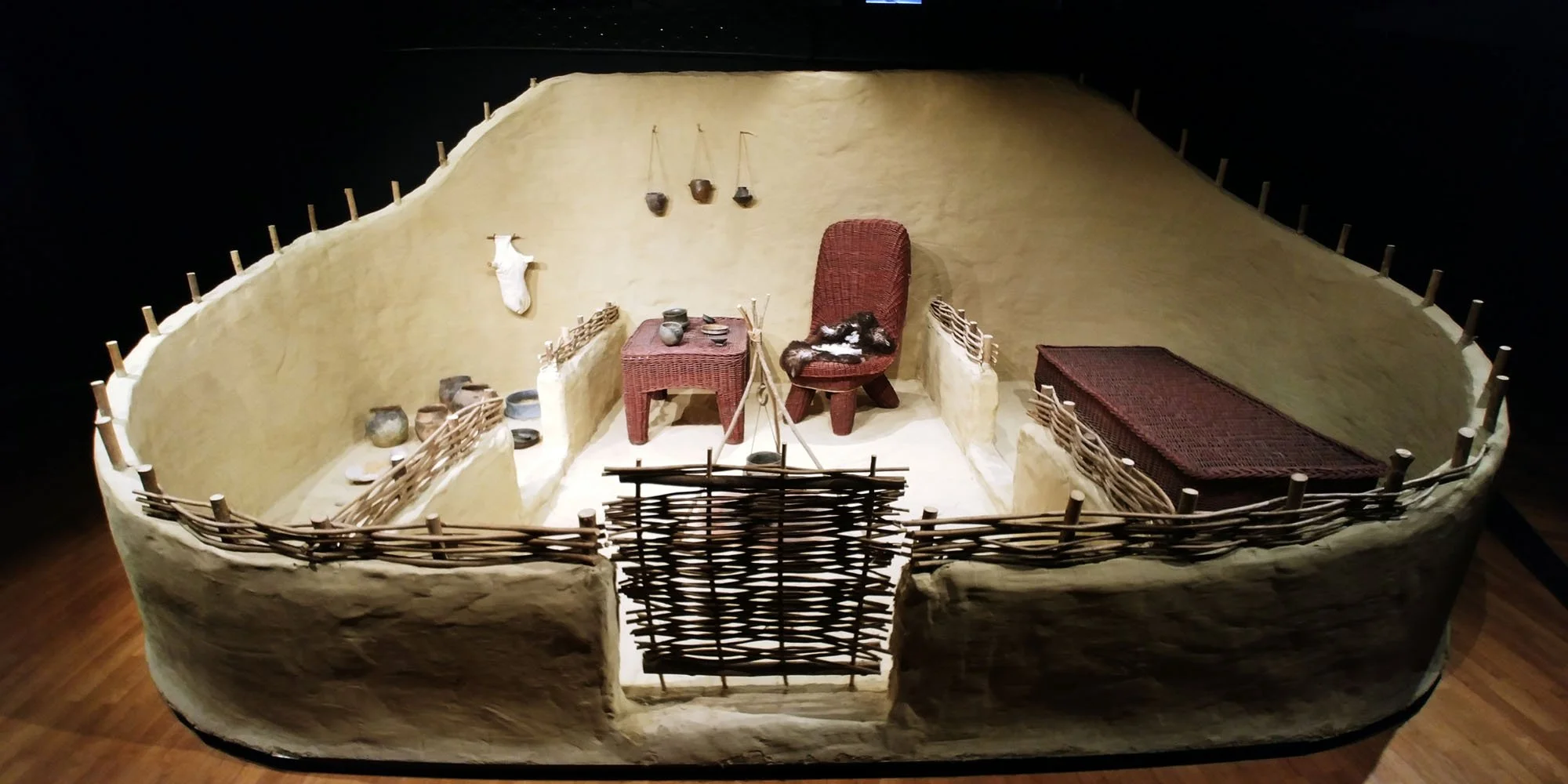 Recreation of a prehistoric or early Native American dwelling interior with pots, a woven bed, and woven fence, viewed from above.