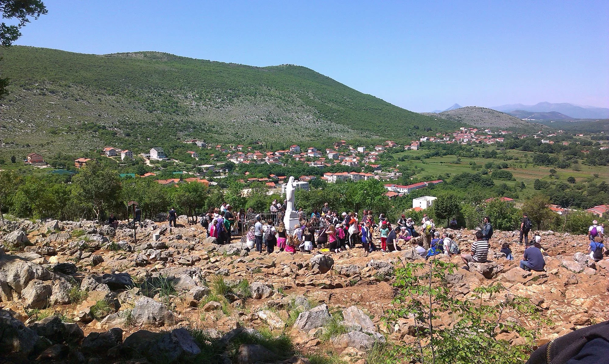 A large group of tourists gathered on rocky terrain at a historical or religious site, with green hills and a small village in the distance under a clear blue sky.