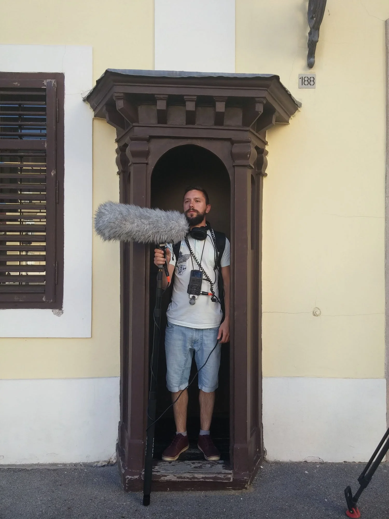 A man holding a boom microphone and wearing headphones stands inside a small wooden porch area in front of a pale yellow building with a window and a plaque marked '188'.