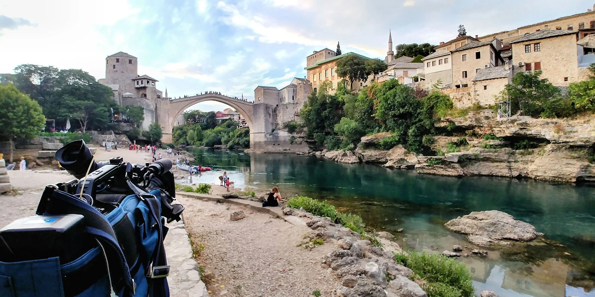 View of a historic stone bridge over a green river, with old buildings and a small minaret on a hillside, and people relaxing on the riverbank in the foreground.