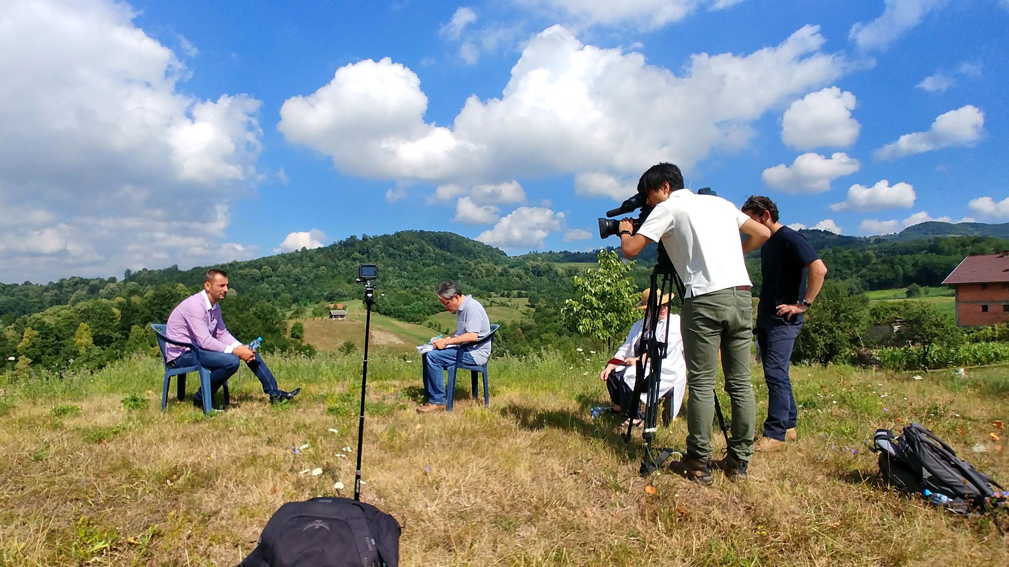 A video crew is recording an interview outdoors in a grassy field with hills and trees in the background. The interviewer and interviewee are seated on chairs, with the crew operating camera and equipment nearby.