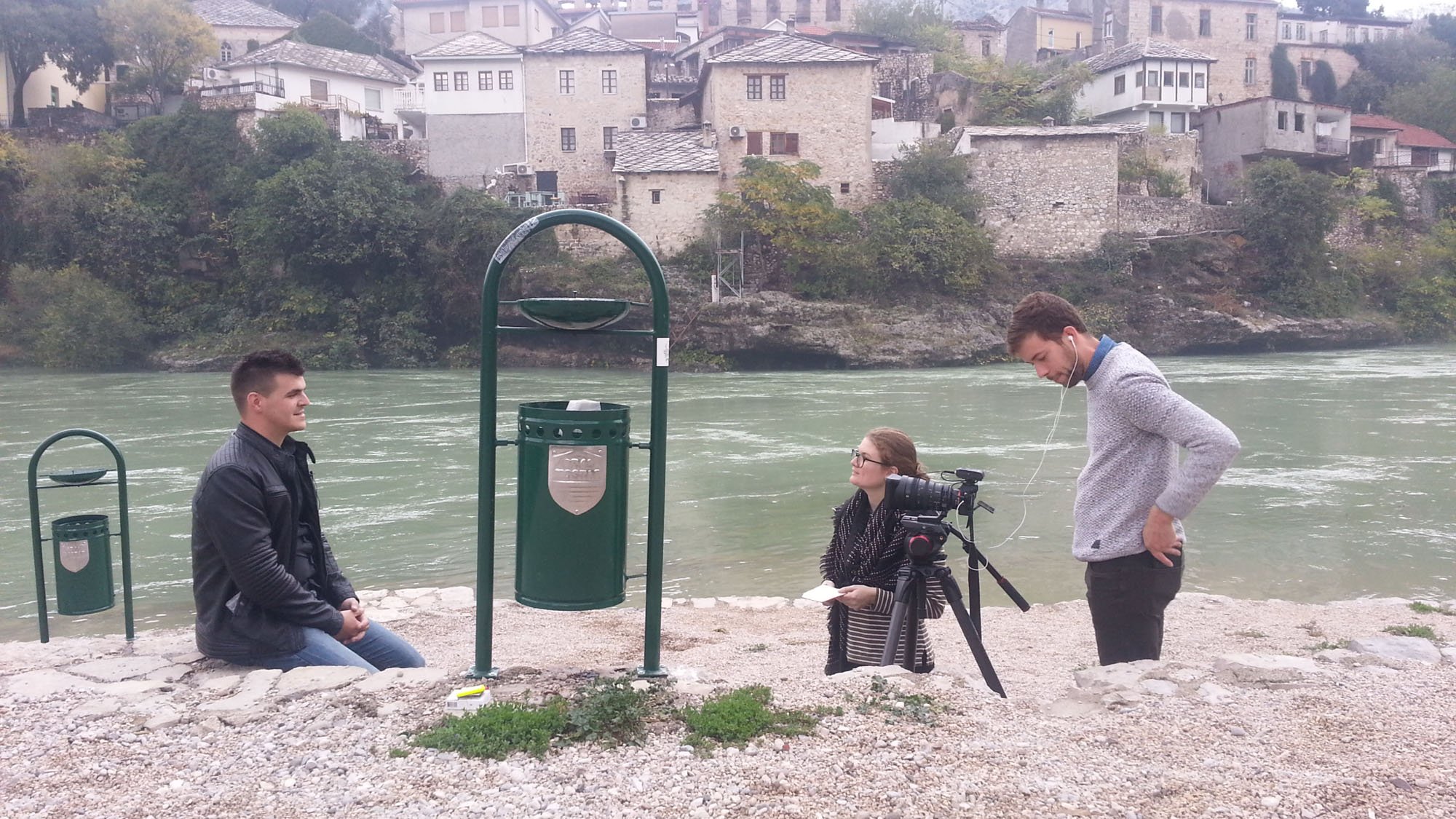 Three young adults beside the river; one man is kneeling, one woman is sitting with a camera, and another man is standing with earphones, in a scenic area with houses on a hill in the background.