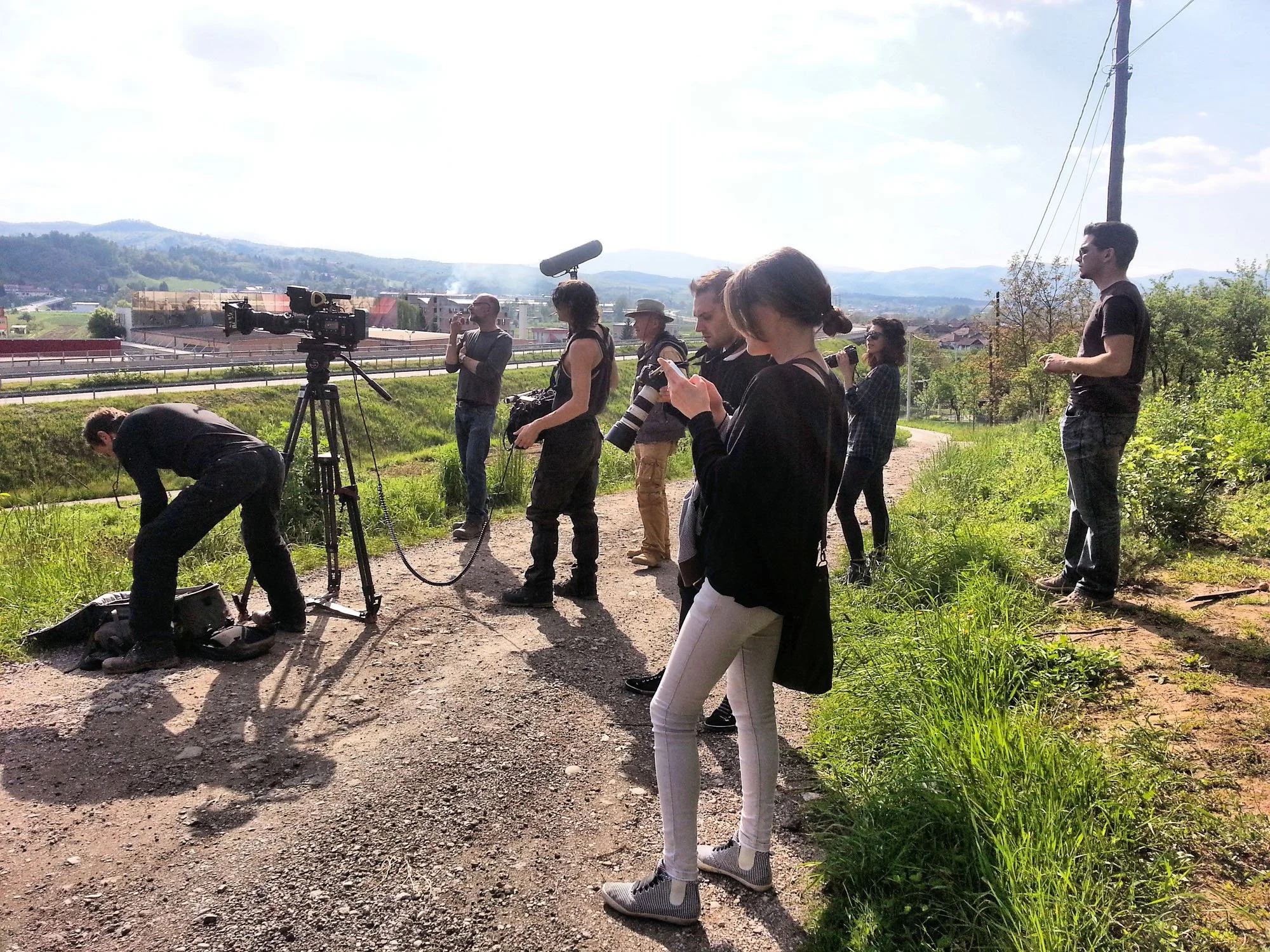 Group of people standing outdoors on a dirt path, some with cameras and equipment, overlooking a rural landscape with rolling hills and utility poles in the background.