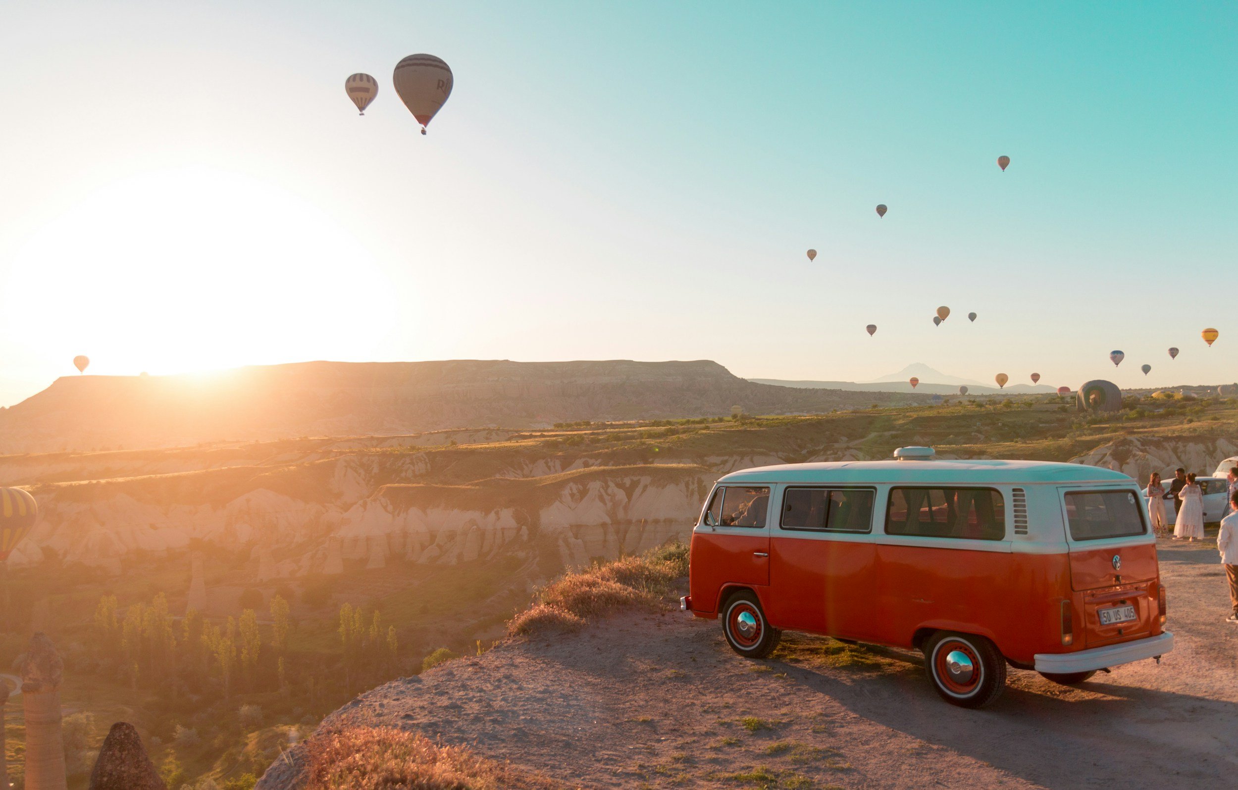 Orange and white retro VW T1 van parked on a cliff with numerous hot air balloons in the sky, illustrating crew transport and recce services by ViEdo in Croatia.