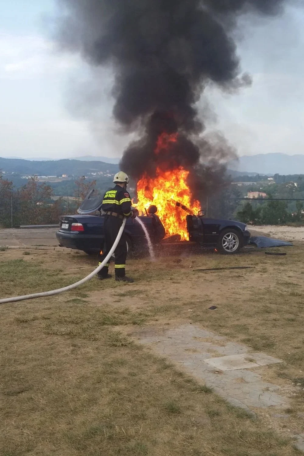 Firefighter extinguishing a car fire on a grassy area, with a background of hills and cloudy sky.