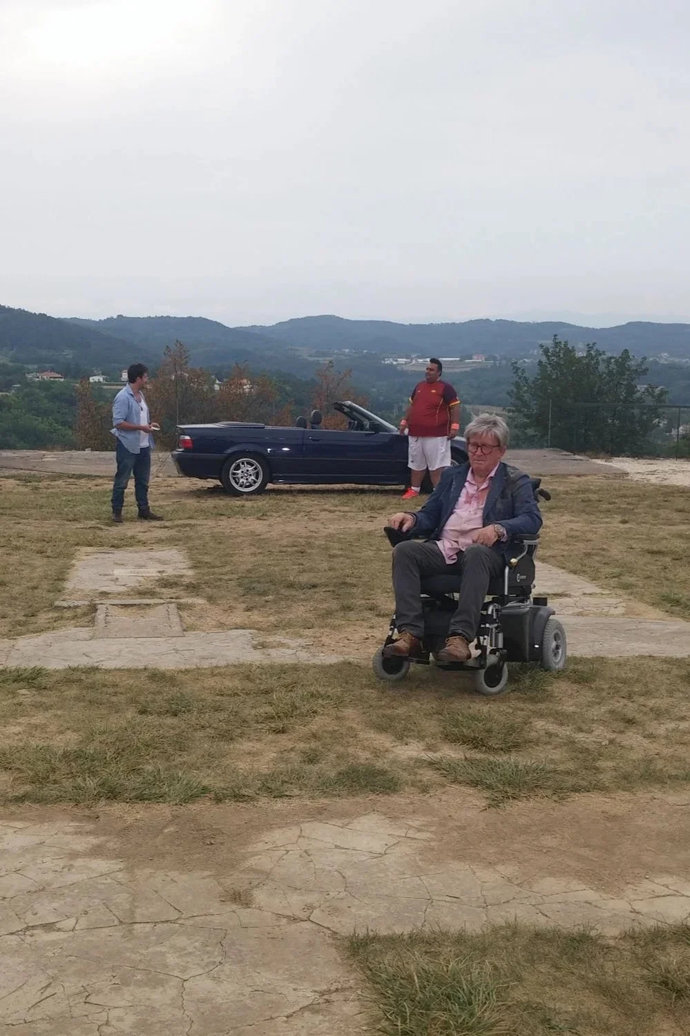 Three men outdoors on a lawn with mountains in the background. One man is sitting in a motorized wheelchair in the foreground, wearing glasses, a pink shirt, and a blazer. Two other men stand near a dark blue convertible car; one on the left wearing 