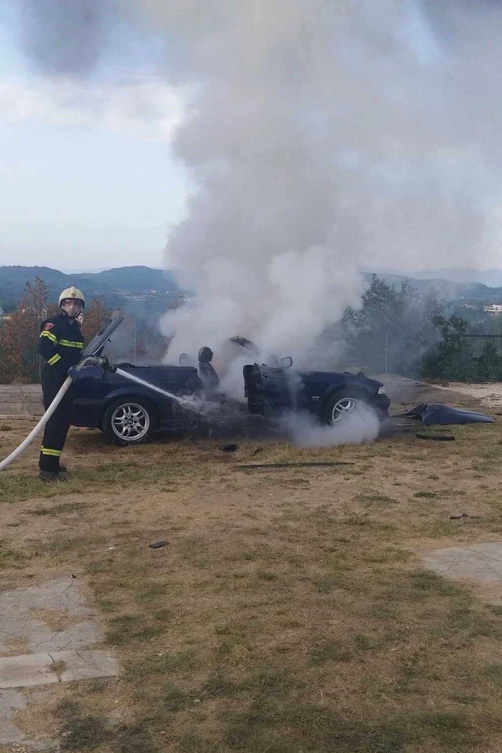 A firefighter in uniform and helmet sprays water on a car that has caught fire, with thick smoke billowing out, in an open area with hills in the background.
