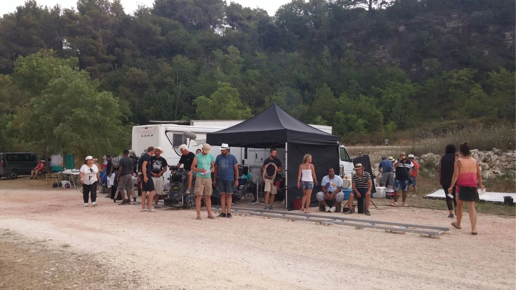 People gathered outdoors around a film set with equipment and a black canopy, with trees and mountains in the background.