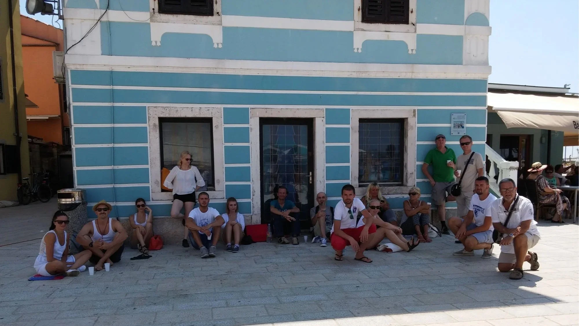 A group of people sitting and standing in front of a blue building during daytime.