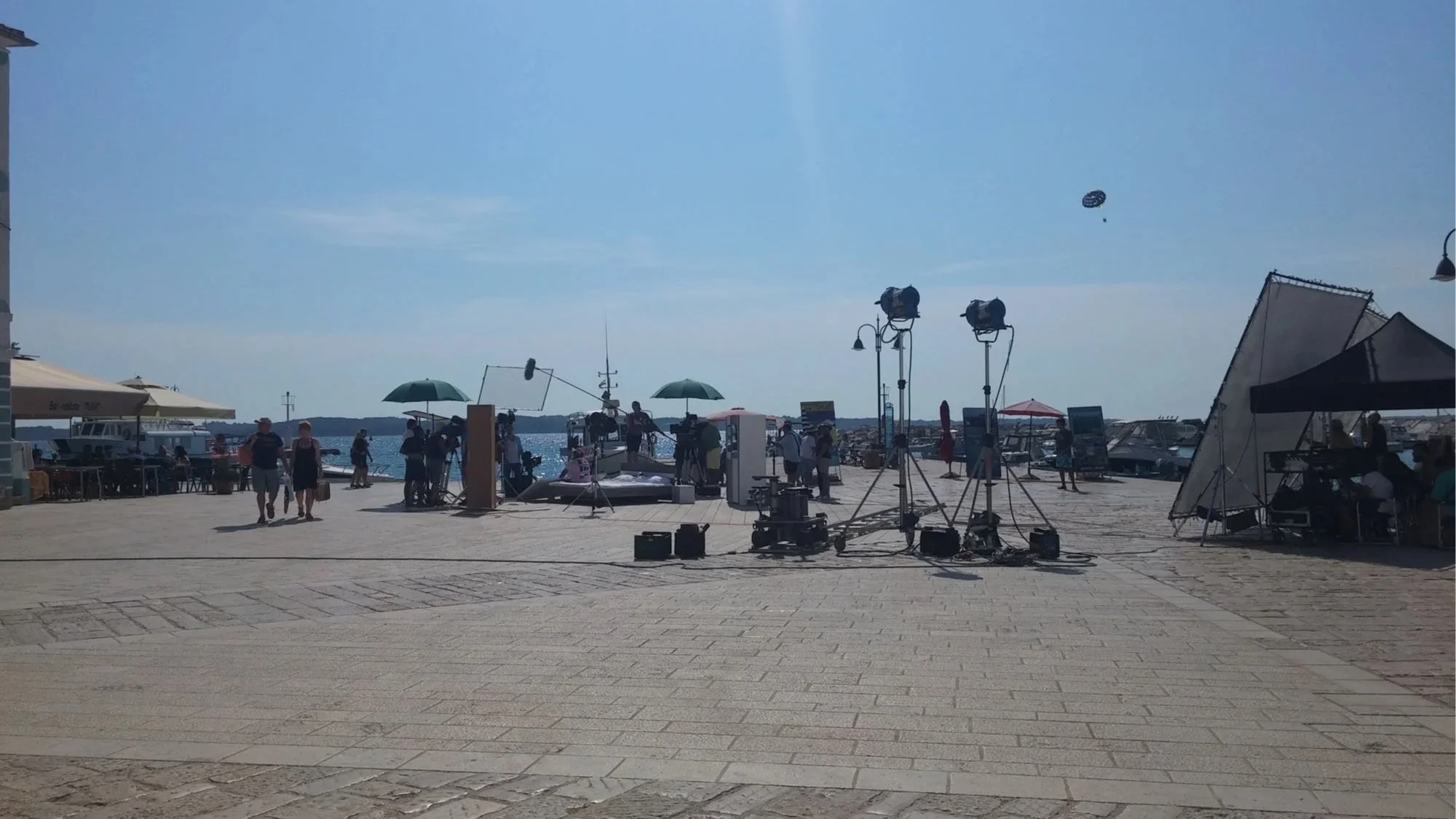 A waterfront scene with film crew equipment and people walking on a paved dock under a clear blue sky, with boats docked in the background and film lights set up.
