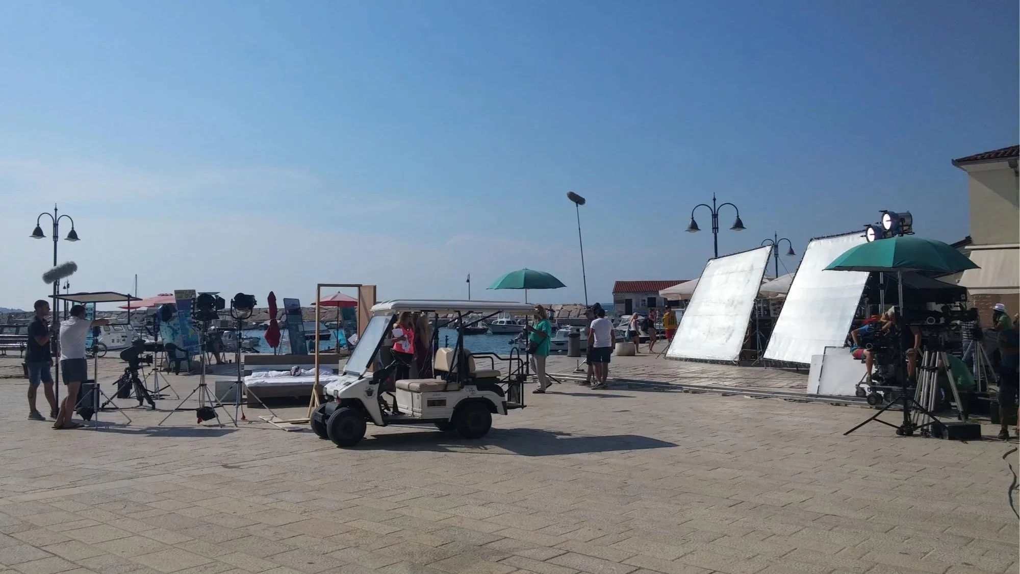 Film crew setting up equipment at a seaside location with boats docked in the background, under a bright blue sky.