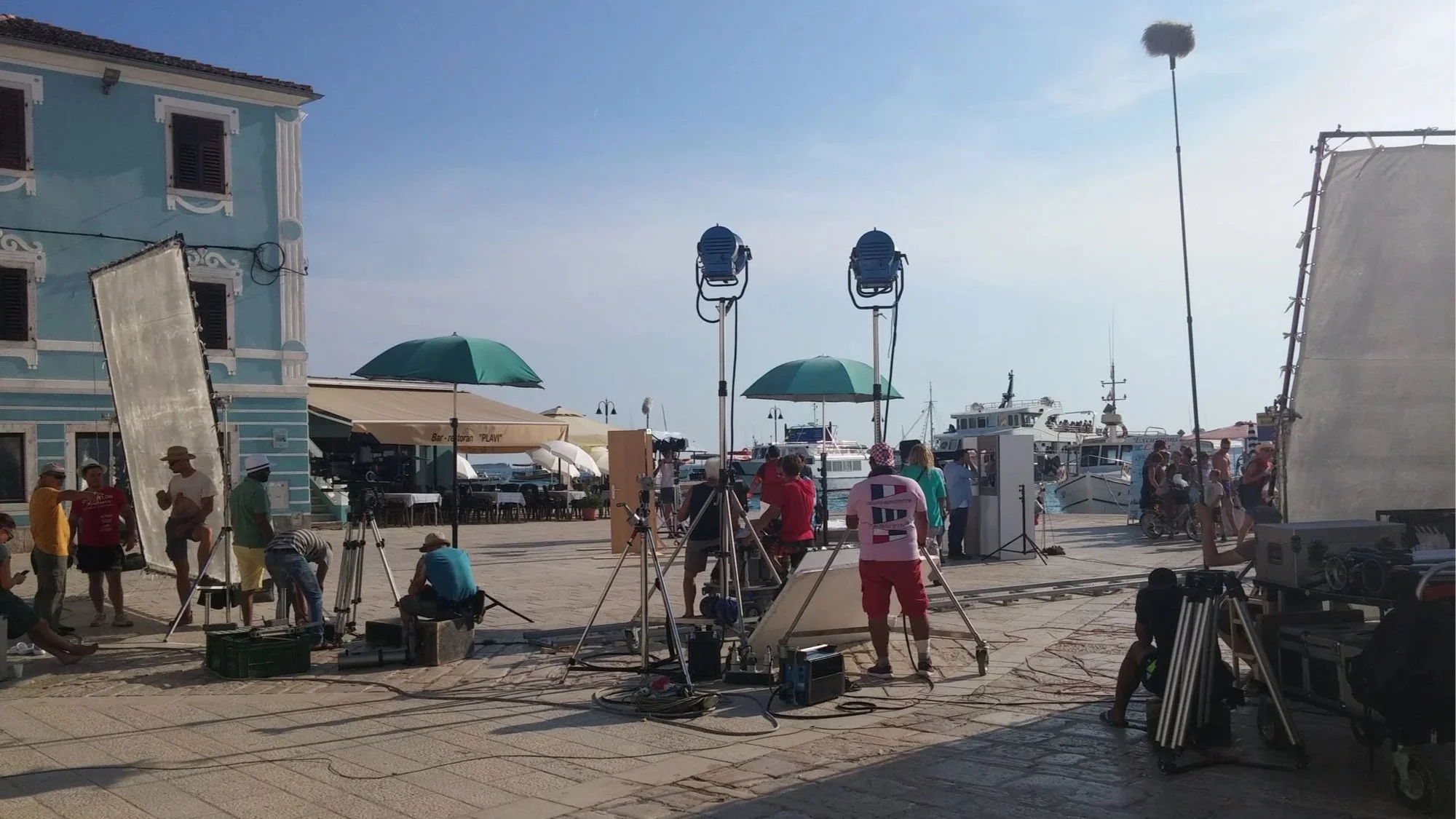 Scene of a film or photo shoot being set up at a harbor with boats docked and people preparing equipment, including lights, tripods, and reflectors, on a sunny day.