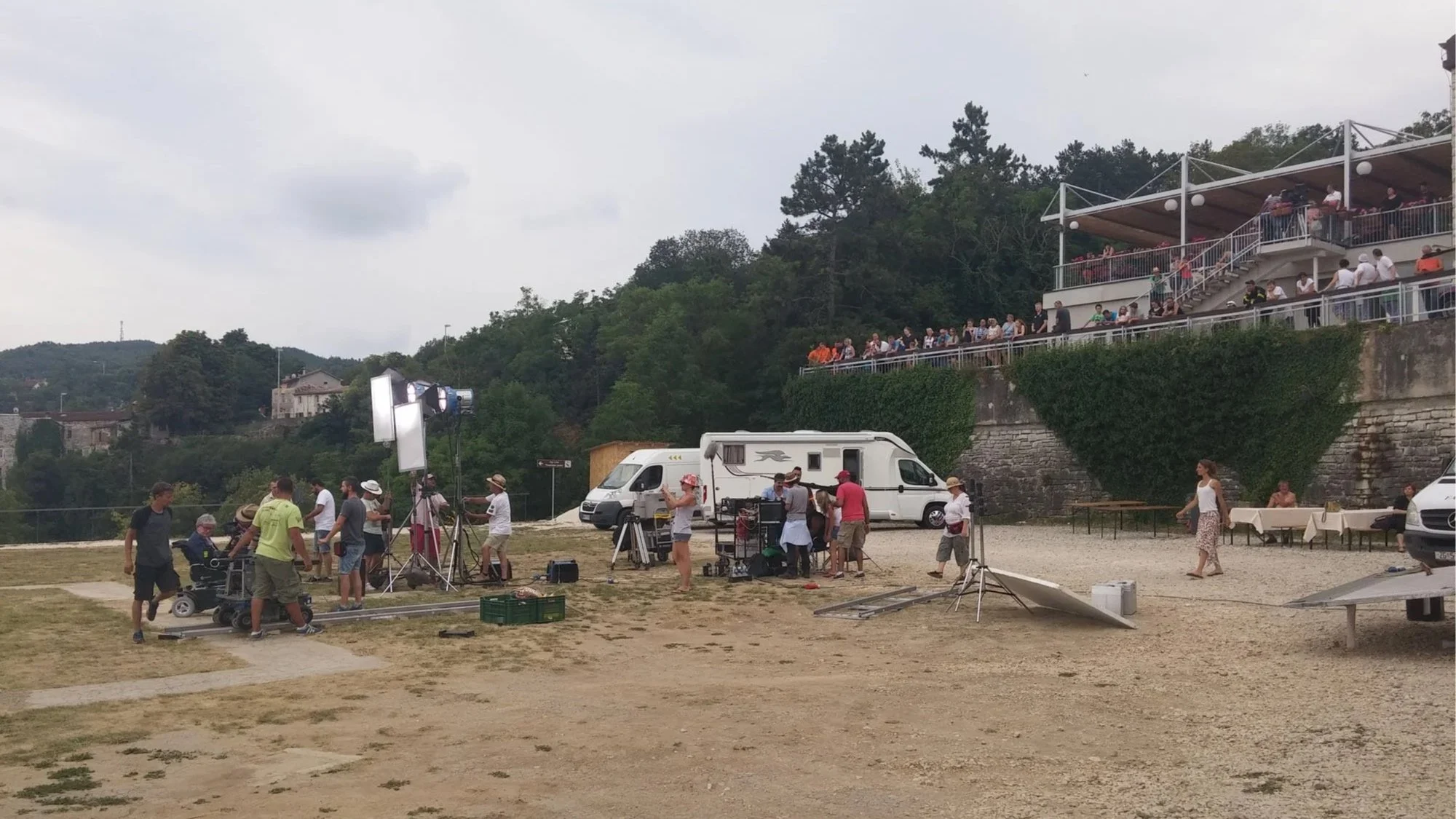 Film crew setting up filming equipment outdoors with a large group of people gathered in a balcony above and others walking around on a sandy ground.
