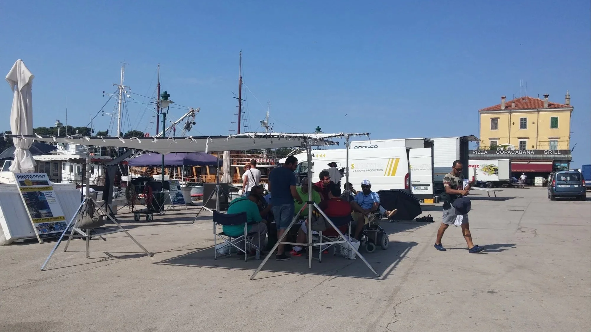 A harbor area with boats, a yellow building with a red roof, and various people sitting and walking around, under a clear blue sky.