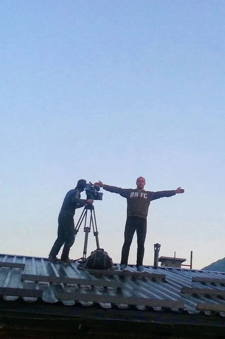 A person standing with arms outstretched on a roof next to a cameraman operating a camera on a tripod, during twilight or early evening.