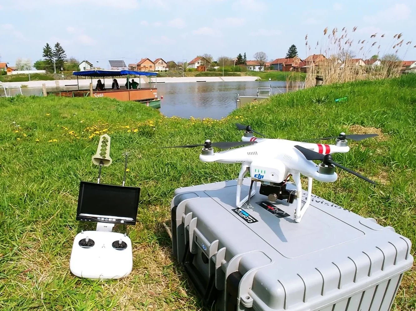 A drone on a gray case with remote control and monitor on grass near a river, with a boat and houses in the background.