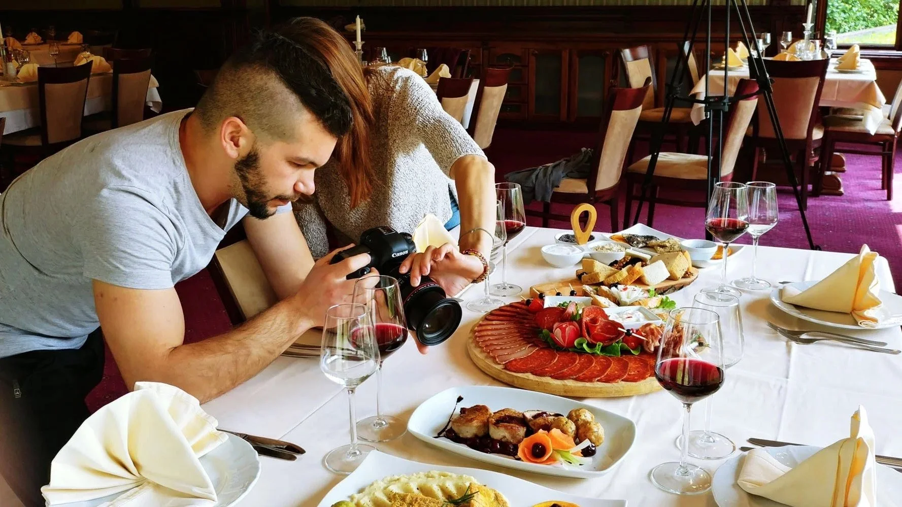 A man taking a photo of a woman at a restaurant table set with wine glasses, plates, and a variety of foods including meats, cheeses, bread, and vegetables.