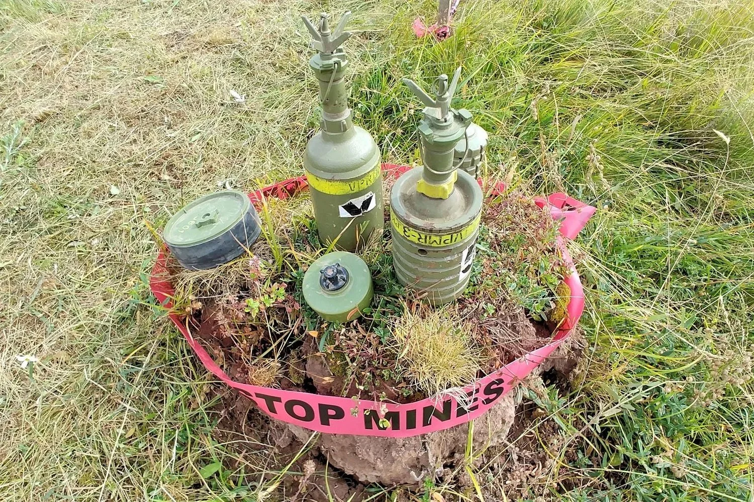 A small area with a pink ring labeled 'STOP MINES' surrounds a cluster of remnants from a landmine, including a pressure-activated device and other metal components, in a grassy outdoor setting with wild plants.