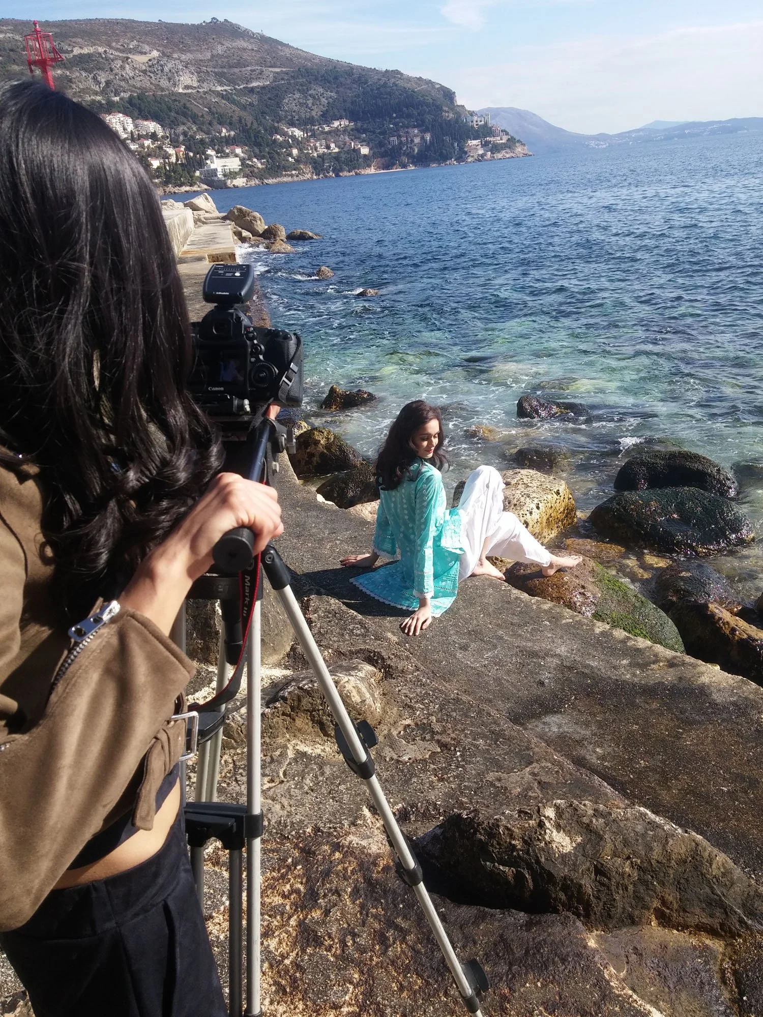 A woman taking photos of another woman sitting on rocks by the water on a coastal area with hills and houses in the background.