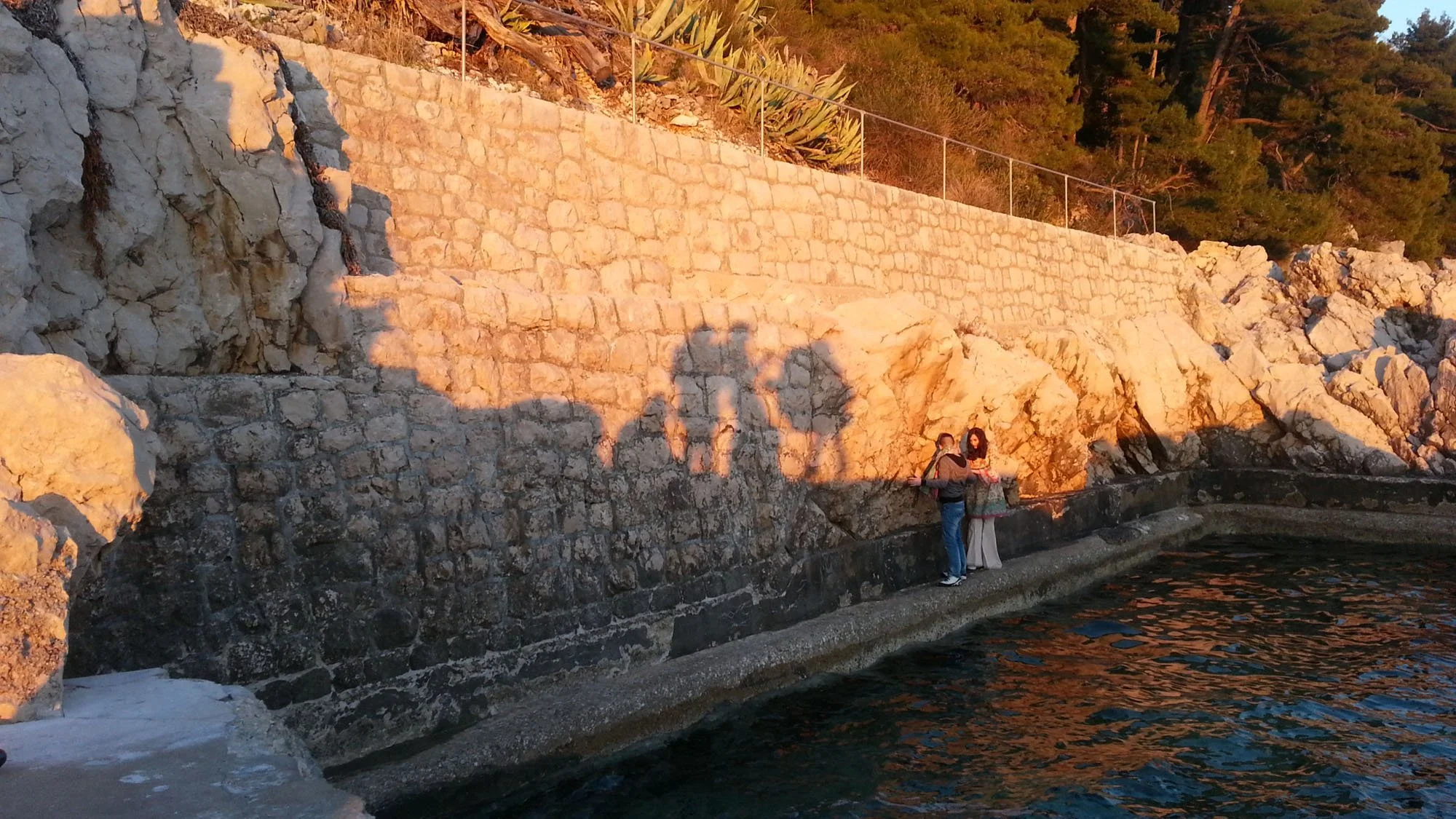 Two people near the water's edge in front of a stone seawall, with a rocky hillside and trees behind them, during sunset.