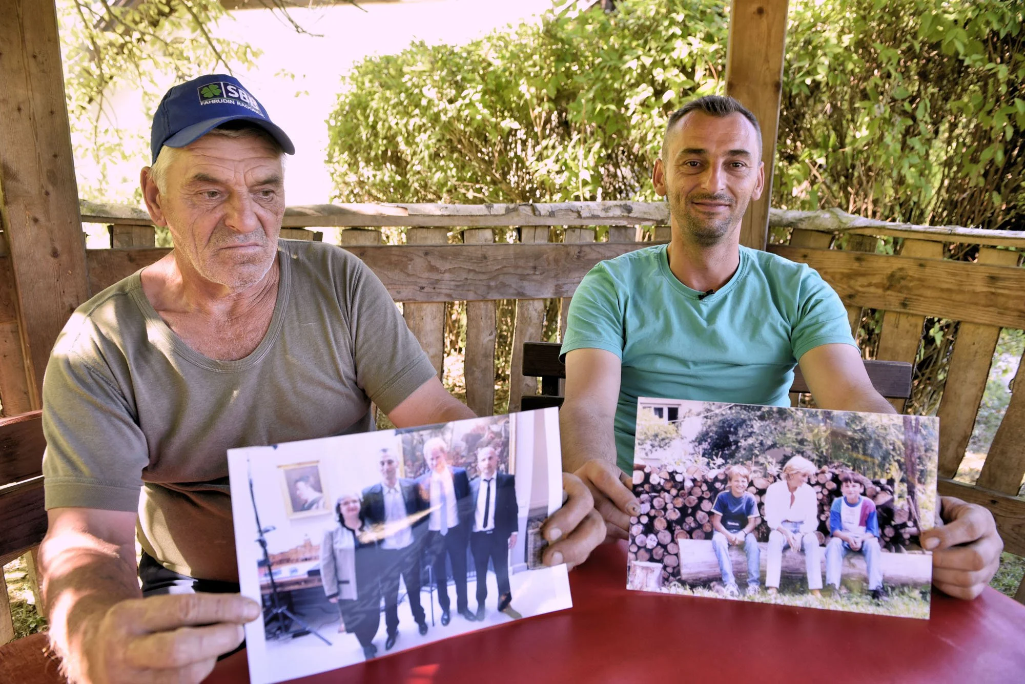 Two men sitting at a wooden outdoor table holding printed photographs of a wedding and children. The older man on the left wears a blue cap, and the younger man on the right wears a turquoise shirt. The background contains greenery and a wooden roof.